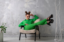 Slender Stylish Young Woman On Heels And Green Suit Sits On Chair On Gray Wall Background In Studio.