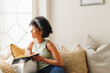 © Wavebreak Media - Thoughtful biracial young woman with afro hair looking through window while writing in diary on sofa