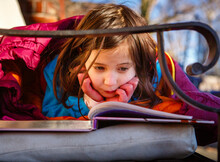 A Little Girl Rests Head On Hands And Reads Book Outside