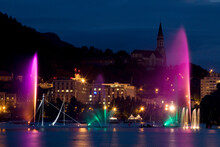 Colored Jets Of Water Burst Upward Rising From A Lake Infront Of A Large Church At Night In Annecy, France.