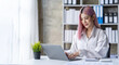 © crizzystudio - Portrait of a young Asian businesswoman sitting at a desk in an office recording data on a laptop. financial calculation and online delivery orders.