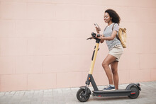 Young Black Woman With Electric Scooter Using Smartphone In A City. Smiling Girl Looking At Mobile Phone On A Street. Active Lifestyle, Eco Transport, Mobile Apps And Online Communication Concept
