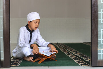 Muslim kid using white rob and skullcap is sitting and reciting Al Quran at the mosque. 