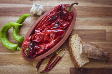 Roasted red peppers in a earthenware pot on a wooden table, accompanied by two slices of traditional natural wheat bread, hot red peppers, shelled walnuts, a head of garlic. Healthy eating, diet