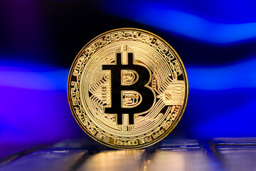 Golden metallic Bitcoin coin standing on a laptop keyboard against the background of a blurred  multicolored sky 