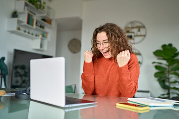 Happy girl student winner looking at laptop receiving good news in email celebrating achievement success. Excited woman winning online, getting new approved job opportunity using computer at home.
