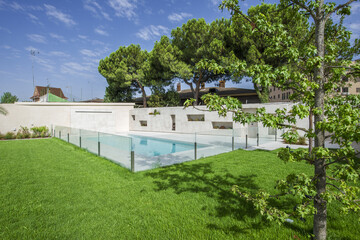 Garden of a detached house with grass and trees and a pool with a glass fence