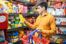 Young Indian Man Buying Grocery At Shop Or Supermarket. Asian Male Choose Snacks And Food Items While Holding Basket.