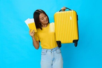 Asian woman holding suitcase and passport with plane ticket smile with teeth in yellow t-shirt and blue jeans on blue background, copy space