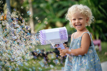Portrait Of A Child Of A Curly Blonde Girl Happily Playing With Soap Bubbles From A Large Air Gun. Outdoors In Summer.