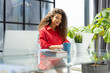© ty - Attractive cheerful business woman in red shirt working on laptop at modern office.
