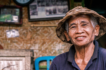 Horizontal portrait of a South Asian seior men wearing a traditional Balinese cone-shaped hat. An elderly man looks into the camera smiling.