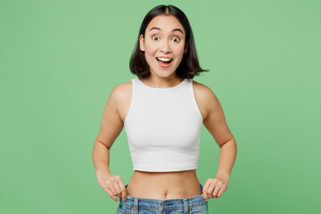 Young excited overjoyed woman wear white clothes show loose pants on waist after weightloss isolated on plain pastel light green background Proper nutrition healthy fast food unhealthy choice concept