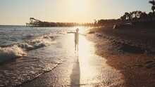 Silhouette Baby Boy Walks Barefoot Along Seashore In Water Waves With Foam Leaving Footprints On Beach Sand. Travel On Landscape Outdoor Nature On Summer Holiday. Children Walk At Sun Rays On Sunset.