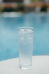 Glass of cold water with ice cubes on table by swimming pool.