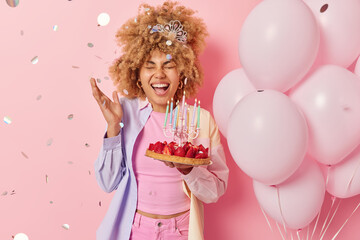 Canvas Print - Happy birthday girl poses with delicious strawberry cake exclaims loudly has fun on party wears crown and colorful shirt stands near bunch of inflated balloons against pink background confetti around