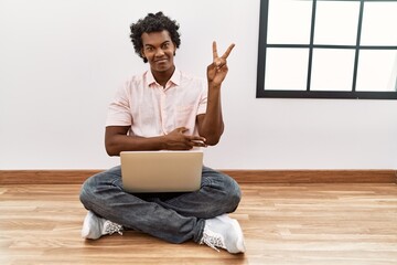 Wall Mural - African man with curly hair using laptop sitting on the floor smiling with happy face winking at the camera doing victory sign. number two.