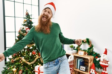 Wall Mural - Redhead man with long beard wearing christmas hat by christmas tree dancing happy and cheerful, smiling moving casual and confident listening to music