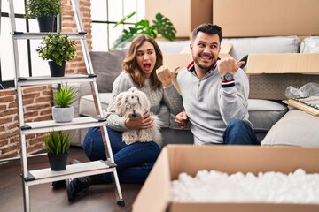 Poster - Young hispanic couple sitting on the floor at new home with log pointing thumb up to the side smiling happy with open mouth