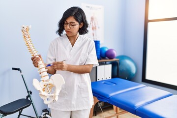 Canvas Print - Young latin woman wearing physiotherapist uniform holding anatomical model of vertebral column at physiotherapy clinic