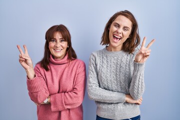 Sticker - Mother and daughter standing over blue background smiling with happy face winking at the camera doing victory sign with fingers. number two.