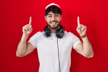 Canvas Print - Hispanic man with beard wearing gamer hat and headphones smiling amazed and surprised and pointing up with fingers and raised arms.