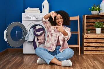 Poster - Young african american woman sitting on the floor doing laundry smiling making frame with hands and fingers with happy face. creativity and photography concept.