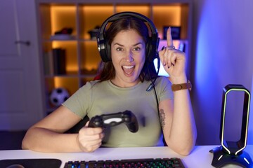 Canvas Print - Beautiful brunette woman playing video games wearing headphones pointing finger up with successful idea. exited and happy. number one.