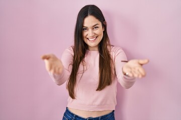 Sticker - Young brunette woman standing over pink background smiling cheerful offering hands giving assistance and acceptance.