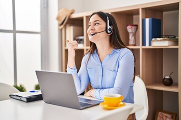 Poster - Young brunette woman wearing call center agent headset smiling with happy face looking and pointing to the side with thumb up.