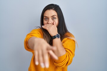 Wall Mural - Young hispanic woman standing over isolated background laughing at you, pointing finger to the camera with hand over mouth, shame expression