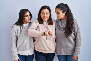 Wall Mural - Mother and two daughters standing over blue background checking the time on wrist watch, relaxed and confident