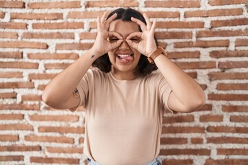 Canvas Print - Young hispanic woman standing over bricks wall doing ok gesture like binoculars sticking tongue out, eyes looking through fingers. crazy expression.