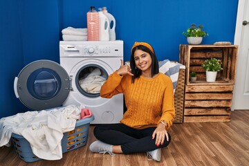 Wall Mural - Young hispanic woman doing laundry smiling doing phone gesture with hand and fingers like talking on the telephone. communicating concepts.