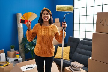 Poster - Young hispanic woman painting home walls with paint roller sticking tongue out happy with funny expression.