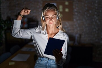 Poster - Young blonde woman working at the office at night strong person showing arm muscle, confident and proud of power