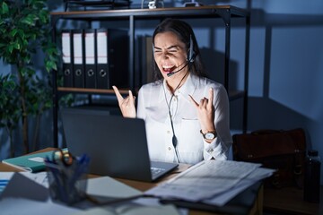 Wall Mural - Young brunette woman wearing call center agent headset working late at night shouting with crazy expression doing rock symbol with hands up. music star. heavy concept.