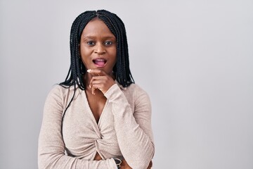 Sticker - African woman with braids standing over white background looking fascinated with disbelief, surprise and amazed expression with hands on chin