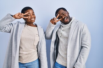 Poster - Young african american couple standing over blue background together doing peace symbol with fingers over face, smiling cheerful showing victory