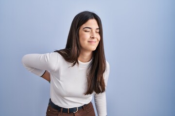 Wall Mural - Young brunette woman standing over blue background suffering of backache, touching back with hand, muscular pain