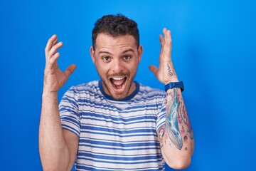 Canvas Print - Young hispanic man standing over blue background celebrating mad and crazy for success with arms raised and closed eyes screaming excited. winner concept