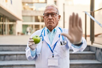 Sticker - Senior doctor with grey hair holding healthy green apple with open hand doing stop sign with serious and confident expression, defense gesture