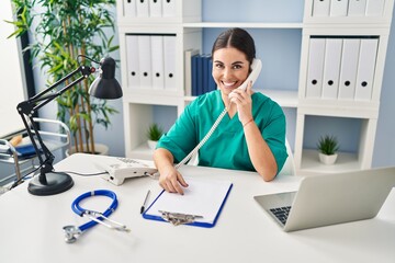 Sticker - Young hispanic doctor woman speaking on the phone at the clinic looking positive and happy standing and smiling with a confident smile showing teeth