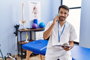 Sticker - Young hispanic man wearing physiotherapist uniform talking on the smartphone at rehab clinic