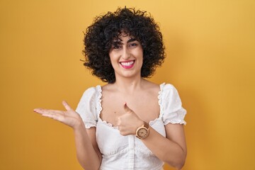 Poster - Young brunette woman with curly hair standing over yellow background showing palm hand and doing ok gesture with thumbs up, smiling happy and cheerful