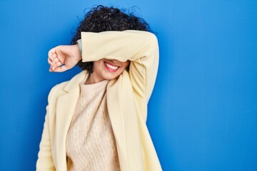 Sticker - Young brunette woman with curly hair standing over blue background covering eyes with arm smiling cheerful and funny. blind concept.