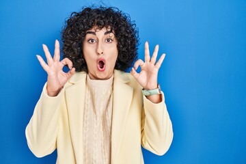 Poster - Young brunette woman with curly hair standing over blue background looking surprised and shocked doing ok approval symbol with fingers. crazy expression