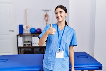 Wall Mural - Young hispanic woman wearing physiotherapist uniform standing at clinic doing happy thumbs up gesture with hand. approving expression looking at the camera showing success.