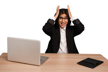 Young indian woman in a table with a laptop and tablet isolated screaming, very excited, passionate, satisfied with something.