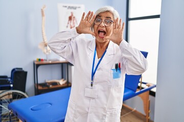 Sticker - Middle age woman with grey hair working at pain recovery clinic smiling cheerful playing peek a boo with hands showing face. surprised and exited